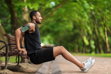 Hot athletic millennial black man exercising in public park, training next to bench, working on arms muscles, using white earbuds, smartphone in armband, listening to music, panorama with copy space