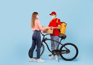 Delivery Offer. Courier Man Delivering Cardboard Box To Lady Customer Standing Wearing Protective Face Mask Over Blue Studio Background. Transportation Service Concept