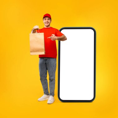Happy Courier Man Showing Paper Bag Near Large Phone Empty Screen Smiling To Camera And Pointing Finger Posing Over Yellow Background. Deliveryman Advertising Delivery Application. Square Shot
