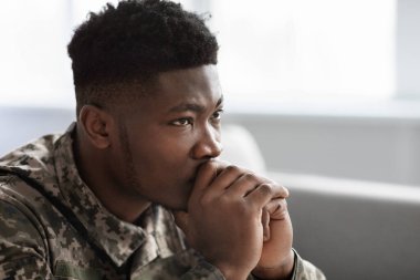 Side view of thoughtful military black man staring at copy space, holding palms by mouth, sitting on couch at home. Soldier visiting psychologist, suffering from posttraumatic stress, closeup