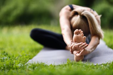 Unrecognizable blonde woman in sportswear stretching body on fitness mat at public park, front view of athletic young lady exercising open-air, doing yoga, copy space