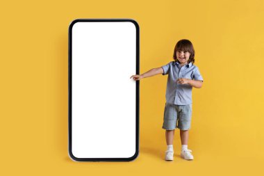 Educational kids mobile app. Happy positive little boy pointing aside at big smartphone with black white screen and smiling to camera, posing over yellow studio background, copy space