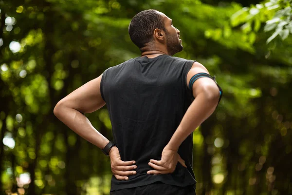 Back view of young black sportsman touching sore zone on his back with both hands, having back pain during jogging by public park, looking at empty space. Workout injuries concept