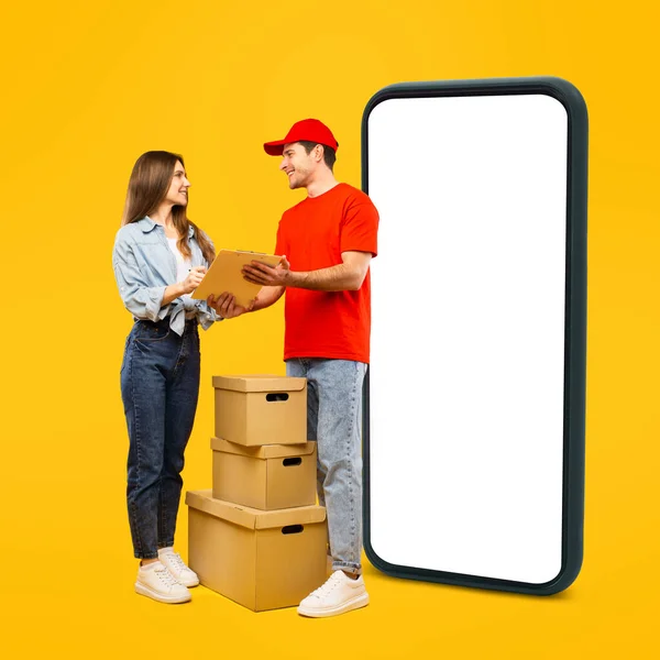 Delivery App. Male Courier And Female Customer Signing Papers Near Big Phone, Deliveryman Delivering Parcel Boxes Standing Over Yellow Studio Background. Mockup, Square Shot