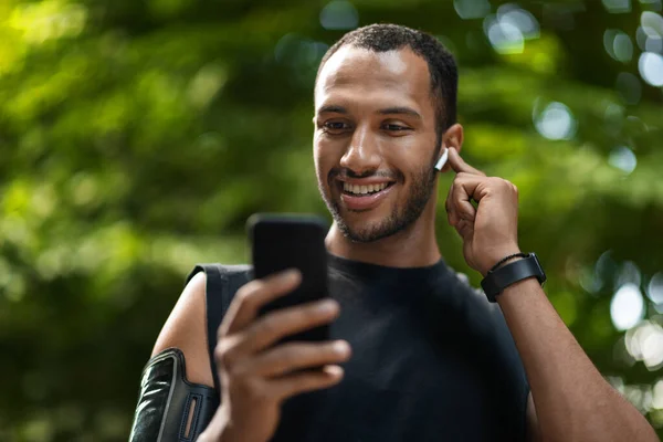 Happy young african american sportsman exercising at public park, using cell phone and wireless earbuds, touching earphone and smiling, having phone call, closeup shot, copy space