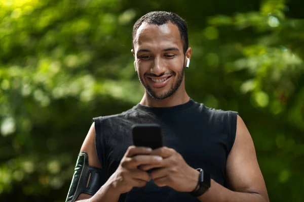 Smiling young black sportsman exercising at public park, using cell phone and wireless earbuds, checking mobile fitness application or setting music for workout, closeup shot, copy space