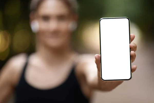 Modern smartphone with white empty screen for advertisement in female hand, unrecognizable sporty woman training outdoors in public park, using fitness mobile app on cell phone, mockup