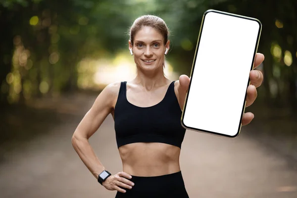 Cheerful young athletic woman in sportswear jogging in park, showing brand new smartphone with white blank screen and smiling, recommending nice mobile fitness app for tracking, mockup,