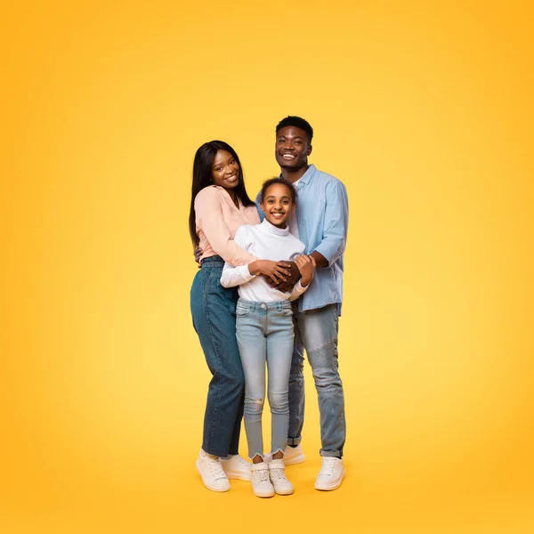 Portrait of happy black family of three embracing and posing together over yellow studio background. Young african american parents hugging their daughter and smiling at camera