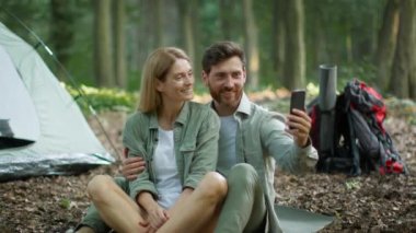 Worldwide communication. Happy active middle aged man and woman video chatting with friends via cellphone app, resting together at forest camping, sitting near tent, slow motion