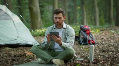 Freelance work comfort. Happy man freelancer working online on digital tablet, sitting near tent at camping, enjoying trip in forest, slow motion, free space