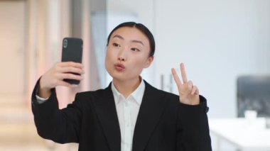 Fun at work. Close up portrait of carefree asian woman manager making selfie on smartphone, showing v sign and grimacing, standing at office, slow motion, free space