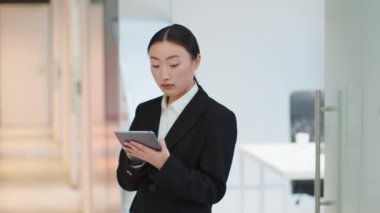 Digital documentation. Concentrated asian lady employee reading emails on digital tablet online, standing at office corridor, slow motion, free space