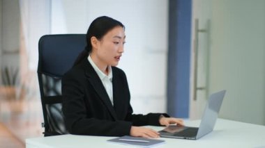 Virtual corporate meeting. Young positive asian woman manager video chatting with colleagues on laptop, waving hand to team, sitting at office, slow motion, empty space