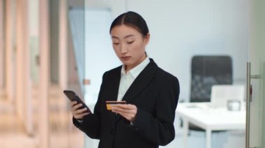 Online payment anywhere. Young asian woman manager making financial transaction with smartphone and credit card, standing at office interior, slow motion, free space