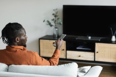 African american man watching television, turning on plasma flatscreen TV-set, pointing remote control at empty TV screen, mockup. Black guy switching channels at home, back view