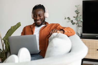Positive black freelancer man in headphones working on laptop at home, sitting on couch in living room, free space. Happy male enjoying remote work