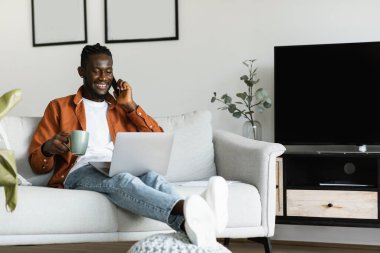 Mobile communication. Cheerful black man using laptop and talking on mobile phone, holding cup and smiling, working remotely while sitting on couch at home interior, copy space