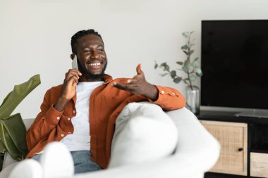 Making call. Happy african american man talking on smartphone and gesturing, resting on sofa at home, free space. Positive male having pleasant conversation
