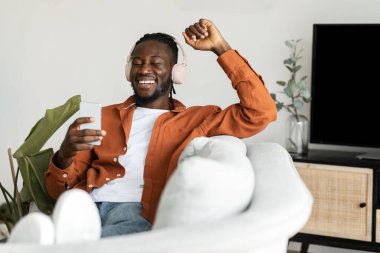 Yes. Overjoyed black man using smartphone and shaking clenched fist, celebrating victory or win, wearing wireless headphones, sitting on the sofa in living room, free space
