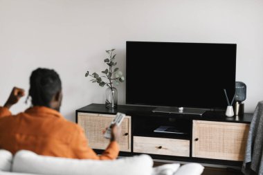 Back view of black man watching sports game on TV, celebrating victory and shaking fists, sitting on sofa in living room. Television set with empty screen. Mockup