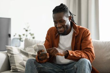 Useful application. Happy africam american man using smatphone while resting on sofa at home, sitting in living room, messaging with friends online