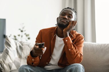 Boring television program. Bored black man watching TV and switching channels with remote control, sitting on couch. Discontented male viewer watching uninteresting movie