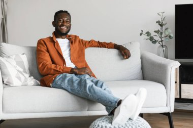Stay at home alone, enjoy silence. Happy african american man sitting on sofa in cozy light living room, looking and smiling at camera, free copy space
