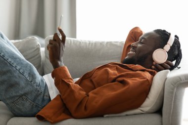 Excited african american man in wireless headphones listening to music on smartphone or watching videos, lying on sofa at home. Happy black guy in headset watching movie on mobile phone