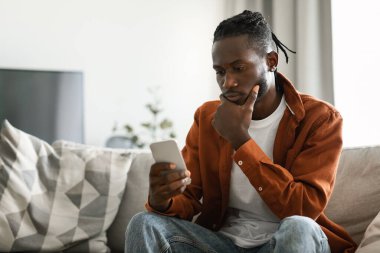 Pensive african american man using smartphone, sitting on sofa at home and looking at phone with serious expression, free space. Black male reading bad news or browsing internet