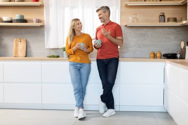Cheerful middle aged married couple having conversation while drinking tea, happy attractive loving man and woman standing by kitchen table, holding coffe mugs and talking, copy space