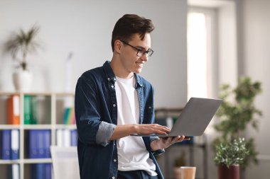 Smiling Millennial Man Using Laptop Computer While Standing Near Desk In Modern Office, Young Male Entrepreneur Wearing Eyeglasses Working Online, Answering Emails, Enjoying Virtual Communication