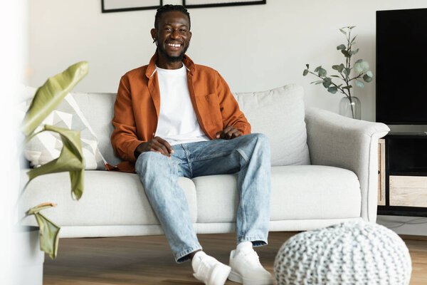 Positive black man sitting on sofa at home, relaxing alone at weekend and smiling at camera, happy african american man posing in cozy living room, full length shot, free space