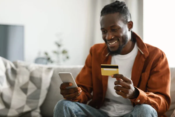 Internet mobile shopping concept. Excited african american man using cellphone and holding credit card, purchasing things online sitting on sofa at home, buying gifts in web store, selective focus