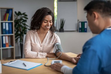 Adult asian man doctor measures pressure of smiling young black female patient in clinic office interior. Examination, hypertension, high blood pressure, treatment of diseases, medical health care