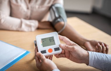 Asian man measures pressure to young black lady patient and shows tonometer screen in clinic. Examination with doctor, hypertension, high blood pressure, treatment disease and medical health care