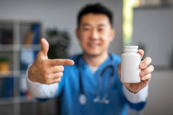 Smiling adult korean male doctor pointing finger at jar of pills in clinic office interior, selective focus. Treatment ill, pharmacy, therapist advice, medical health care during covid-19 pandemic
