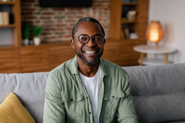 Gözlüklü, rahat bir şekilde kanepede dinlenen ve oturma odasının iç tarafındaki kameraya bakan Afro-Amerikan bir adamın portresi. COVid-19 karantinası sırasında iş ve evde çalışma