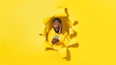 Duygusal şokta olan Afro-Amerikan adam akıllı telefon kullanıyor ve kameraya ağzı açık bakıyor, haberleri okurken yanağına dokunuyor, sarı kağıtta yırtık bir delikte poz veriyor, panorama
