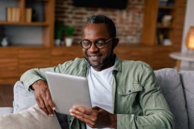 Gözlüklü, neşeli, Afro-Amerikan bir adam. Tablet üzerinde günlük videolar izleyip, oturma odasında sohbet ediyor. Yeni uygulama, iş kumandası, sosyal medyada blog, covid-19 karantinasında yeni normal