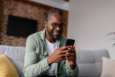 Gözlüklü ve günlük izleme videolu mutlu Afrikalı Amerikalı adam akıllı telefonda sohbet ediyor, oturma odasındaki kanepede dinleniyor. İş ve uzaktan çalışma evde, blog, yeni normal ve yeni uygulama