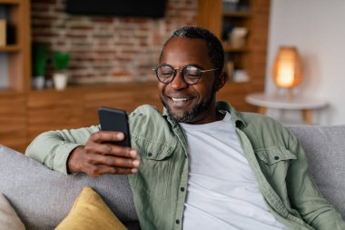 Orta yaşlı neşeli Afro-Amerikan erkek, gözlüklü ve akıllı telefonla sohbet eden oturma odasındaki kanepede dinlenen. Sosyal medya, blog, yeni uygulama, iş ve evde çalışma süresi covid-19