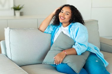 Evdeki kameraya gülümseyen Afro-Amerikan bayan yastık pozisyonunda dinleniyor. Rahatlamış kadın hafta sonları evdeki rahatlığın tadını çıkarıyor. Dinlenme ve Rahatlama