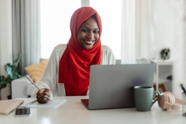 Not alıyorum. Pozitif Müslüman Afro-Amerikan kadın iş yerinde oturuyor, dizüstü bilgisayar kullanıyor ve not defterine yazıyor. Heyecanlı siyahi kadın evden internetten çalışıyor, uzaktan çalışıyor.