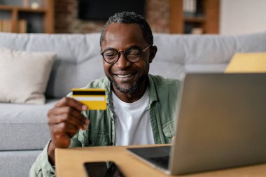 Orta yaşlı gözlüklü Afro-Amerikalı adam oturma odasının içinde kredi kartıyla online alışveriş yapmaktan hoşlanıyor. Evdeki teknoloji, satış ve finans bankacılığı ile yeni normal
