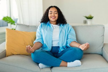 Mutlu Afrika kökenli Amerikalı kadın evde Lotus pozisyonunda otururken gözleri kapalı meditasyon yapıyor. Oturma odasında yoga yapan bayan dinleniyor. Meditasyon ve rahatlama. Seçici Odaklanma