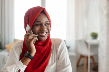 Tesettürlü siyahi kadın cep telefonuyla arama yapıyor, gülümsüyor ve başka tarafa bakıyor, evde oturuyor. Orta Doğulu mutlu bir kadın oturma odasında akıllı telefondan konuşuyor.