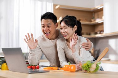 Asyalı eşlerin mutfakta yemek pişirirken arkadaşlarıyla ya da aileleriyle görüntülü konuşma yapmaları, sebze dolu masanın yanında durmaları, dizüstü bilgisayarda el sallamaları ve gülümsemeleri,