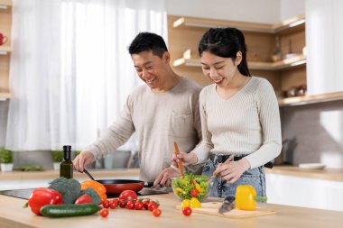 Evde birlikte yemek pişirirken gülümseyen Asyalı eşler, sağlıklı yemek hazırlayan genç eş ve orta yaşlı koca, sohbet eden, kopyalama alanı. Beslenme, sağlıklı beslenme konsepti