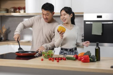 Neşeli Asyalı çift orta yaşlı koca ve genç eş evde birlikte yemek pişirmekten zevk alıyorlar, mutlu Çinli erkek ve kadın sobanın yanında duruyor, sağlıklı bir akşam yemeği hazırlıyorlar, mekanı kopyalıyorlar.